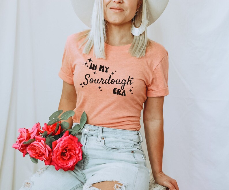 K&ouml;nnte beinhalten: Eine Frau tr&auml;gt ein pfirsichfarbenes T-Shirt mit dem Text "In My Sourdough Era" in schwarzen Buchstaben. Sie tr&auml;gt einen wei&szlig;en Hut und eine blaue Jeans.