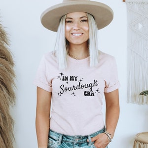 K&ouml;nnte beinhalten: Ein rosa T-Shirt mit dem Text "In My Sourdough Era" in schwarzer Schrift mit kleinen Sternen um den Text.