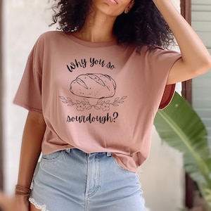 Peut inclure: Un t-shirt rose p&acirc;le avec un graphique noir d'une miche de pain et le texte "Why you so sourdough?"