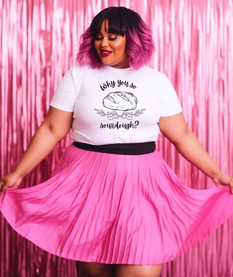 Peut inclure: Un t-shirt blanc avec un motif noir repr&eacute;sentant un pain et le texte "Why you so sourdough?" Le mannequin porte une jupe pliss&eacute;e rose et a les cheveux roses.