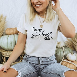 K&ouml;nnte beinhalten: Eine Frau tr&auml;gt ein wei&szlig;es T-Shirt mit dem Text "In My Sourdough Era" in schwarzen Buchstaben. Sie tr&auml;gt eine blaue Jeans und eine goldene Halskette.