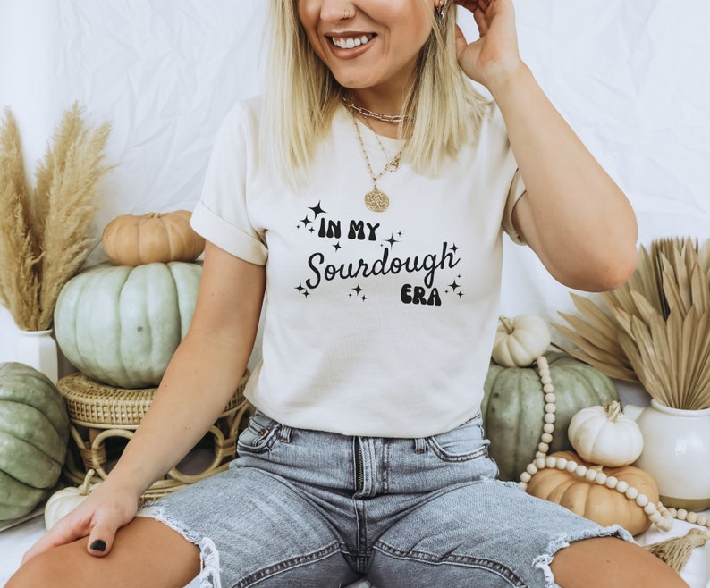 K&ouml;nnte beinhalten: Eine Frau tr&auml;gt ein wei&szlig;es T-Shirt mit dem Text "In My Sourdough Era" in schwarzen Buchstaben. Sie tr&auml;gt eine blaue Jeans und eine goldene Halskette.