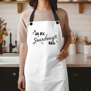 K&ouml;nnte beinhalten: Wei&szlig;e Sch&uuml;rze mit schwarzem Text, der "In My Sourdough Era" lautet, mit Sternen um den Text herum. Die Sch&uuml;rze hat verstellbare schwarze Tr&auml;ger.
