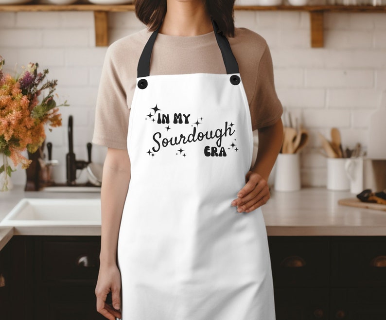 K&ouml;nnte beinhalten: Wei&szlig;e Sch&uuml;rze mit schwarzem Text, der "In My Sourdough Era" lautet, mit Sternen um den Text herum. Die Sch&uuml;rze hat verstellbare schwarze Tr&auml;ger.