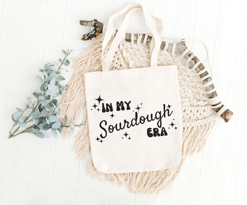 K&ouml;nnte beinhalten: Eine wei&szlig;e Stofftasche mit schwarzem Text, der "In My Sourdough Era" lautet, mit funkelnden Sternen um den Text herum.