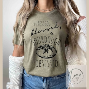 May include: A green t-shirt with the text "Stressed, Blessed, & Sourdough Obsessed."  A black and white illustration of a loaf of sourdough bread is below the text.
