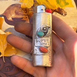 May include: Silver lighter with turquoise and red stone accents, and the words "SUPER SHIT" engraved on the front. The lighter has a decorative design and a red button.
