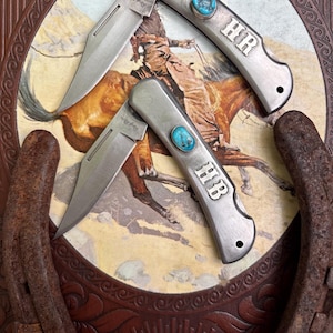 May include: Two silver pocket knives with turquoise accents and the initials "HR" and "HB" engraved on the handles. The knives are displayed on a decorative background featuring a cowboy on horseback and a horseshoe.