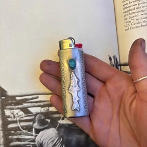 May include: A silver lighter with a turquoise stone and a fish design. The lighter has a red pull tab and is held in a hand. The background includes a book and a black and white photograph.