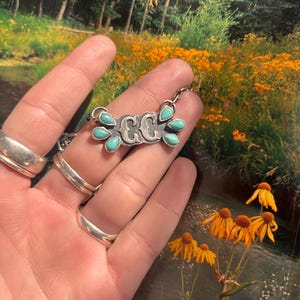 May include: A silver necklace with a pendant featuring the initials "CC" and turquoise teardrop-shaped stones. The necklace is held up against a backdrop of a natural landscape with yellow flowers and a stream.