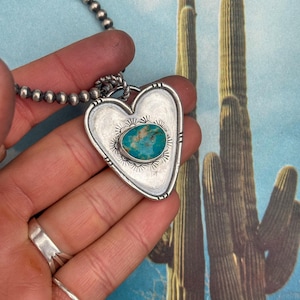 Op de afbeelding: Zilveren hartvormige hangerketting met een turquoise steen. De hanger heeft een gedetailleerd, getextureerd ontwerp en hangt aan een kralenketting. De achtergrond toont een woestijnscène met cactussen.