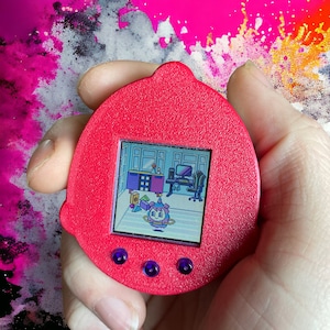 May include: A red handheld digital pet device with a screen showing a cartoon character. The screen is surrounded by a black border and has three purple buttons below it.