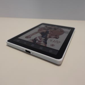 May include: A white rectangular electronic device with a black screen displaying a graphic illustration of a person, the number 4,122, and various icons. The device features a centered charging port on the bottom edge and a thin black bezel.