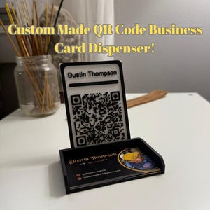 May include: A custom-made black QR code business card dispenser. The dispenser has a clear acrylic top with the name "Dustin Thompson" and a QR code. The base holds business cards with the name "Dustin Thompson" and contact information.