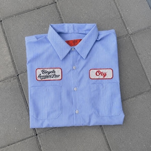 May include: A light blue and white pinstriped button-down work shirt featuring a pointed collar and two front pockets. Two rectangular patches are sewn onto the chest; the left reads Bicycle Accident Law and the right reads Ory in red script.