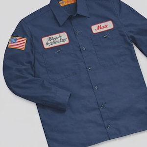 May include: A navy blue long-sleeve button-down work shirt featuring a collar, two chest pockets, and a button-up front. Patches include a United States flag on the right sleeve, a "Bicycle Accident Law" patch, and a "Matt" name patch on the chest.