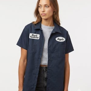 May include: A navy blue short-sleeved button-down work shirt featuring two chest pockets. A white rectangular patch on the right side reads Bicycle Accident Law, and a white oval patch on the left side features the name Angela in black script.
