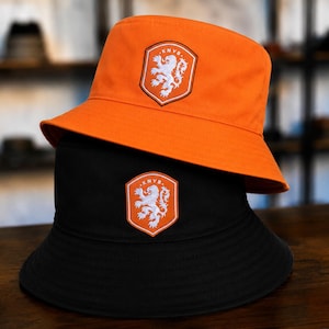 May include: An orange bucket hat stacked on top of a black bucket hat. Each hat features a centered shield-shaped patch with a white lion emblem and the text KNVB embroidered in white. The hats have a wide, downward-sloping brim and a flat crown.