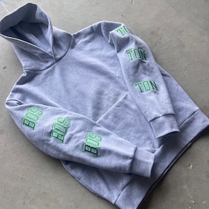 Puede incluir: Una sudadera con capucha gris con letras bordadas en verde en las mangas. Las letras de la manga izquierda dicen "BOS" tres veces, y las letras de la manga derecha dicen "TON" tres veces.
