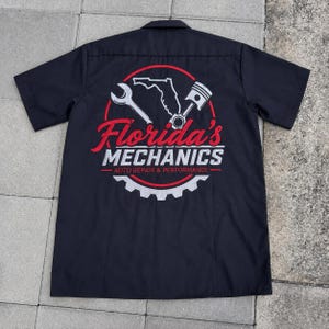 May include: A black short-sleeve button-down shirt featuring a graphic on the back. The design includes a red circle, a wrench, a piston, and the outline of Florida. Text reads Florida's MECHANICS AUTO REPAIR & PERFORMANCE in red and white.