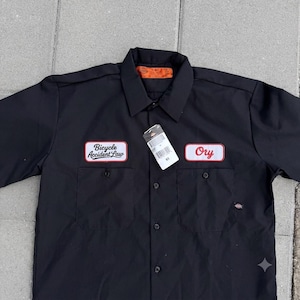 Pode incluir: Uma camisa de trabalho preta de manga curta com colarinho, fecho de botões e dois bolsos no peito. Apresenta dois emblemas retangulares bordados: um com o texto Bicycle Accident Law e outro com Ory. Uma etiqueta branca pende do colarinho.