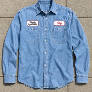 May include: A light blue denim long-sleeve button-down shirt featuring two chest pockets and two rectangular embroidered patches. The left patch reads Bicycle Accident Law in black text, and the right patch reads Ory in red cursive script.