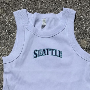 May include: A light blue ribbed tank top with the word "SEATTLE" arched across the front in teal lettering. The tank top has a rounded neckline and wide shoulder straps. The fabric appears to be a soft, comfortable material.