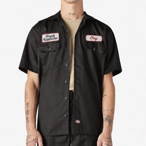 May include: A black short-sleeved button-down work shirt featuring two chest pockets with flaps. The left pocket has a white patch with red trim reading Bicycle Accident Law, and the right pocket has a white patch with red trim reading Ory.