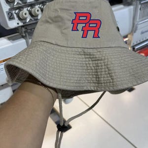 May include: A beige bucket hat with a red and blue embroidered "PR" logo.