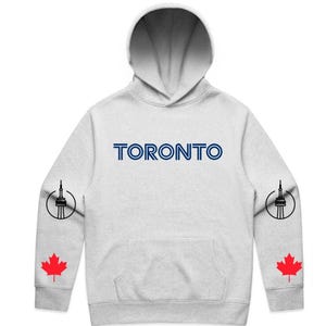 Puede incluir: Sudadera con capucha gris claro con la palabra "TORONTO" en letras azules en el pecho. Las mangas presentan una ilustración negra de la Torre CN y una hoja de arce roja.