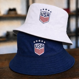 Pode incluir: Um chapéu bucket branco sobreposto a um chapéu bucket azul-marinho. Ambos os chapéus apresentam um emblema bordado na frente com o texto USA, cinco estrelas acima do escudo e listras verticais vermelhas e brancas no design.