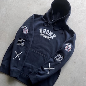 Puede incluir: Una sudadera con capucha azul marino con el texto "Bronx Bombers" bordado en la parte delantera. Las mangas tienen bordados de bates de béisbol, pelotas de béisbol y las letras "BX".