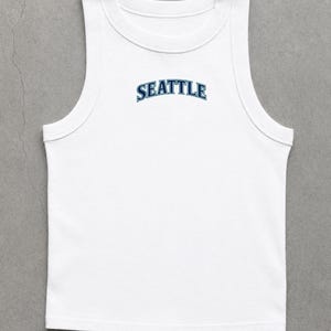 May include: White tank top with a rounded neckline and a sleeveless design. The word "SEATTLE" is arched across the chest in blue lettering.