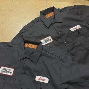 May include: Two charcoal gray short-sleeve work shirts featuring a button-down front, a pointed collar, and two chest pockets. Each shirt displays a white rectangular patch with the red text Bicycle Accident Law and a name tag patch.