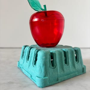 May include: A translucent red apple-shaped container with a green leaf accent on top, resting on a square, light teal-colored molded pulp berry basket with vertical ventilation slots along its four sides.