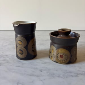May include: A dark brown ceramic creamer and a matching lidded sugar bowl. Both pieces feature a repeating pattern of large, circular, mustard-yellow and tan motifs with small, dark brown centers. The creamer has a white interior and a spout.