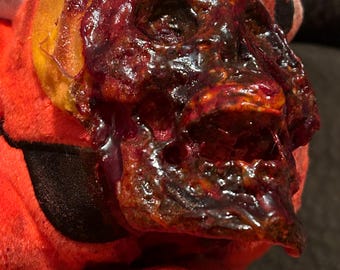 May include: A close-up of a red and orange plush toy with a bloody, melted skull face. The skull is dripping with red and brown goo.