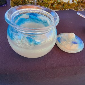 May include: A clear glass jar with a lid, featuring a blue and white swirled design. The jar has a wide opening and a rounded shape. The lid is off to the side, showing the same blue and white pattern. The jar sits on a dark surface.