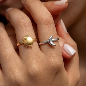 Puede incluir: Dos anillos, uno dorado con un diseño de sol y otro plateado con una media luna. El anillo solar tiene un centro redondo y texturizado con rayos puntiagudos. El anillo lunar tiene una forma simple y curvada. Ambos anillos están en un dedo.