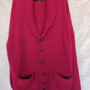 Könnte beinhalten: Ein dunkelroter Cardigan mit einem breiten Kragen, fünf schwarzen Knöpfen und zwei aufgesetzten Taschen.