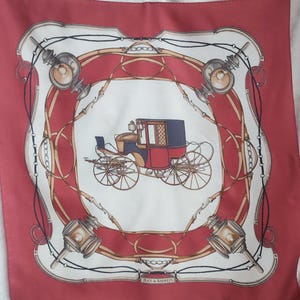 Pode incluir: Um lenço de seda vermelho com um centro branco que apresenta uma carruagem puxada por cavalos preto e vermelho com detalhes dourados. A borda do lenço é um padrão repetido de correntes douradas, correias de couro marrom e lanternas douradas. O texto "JEAN de BAHREIN" está impresso em dourado na parte inferior do lenço.
