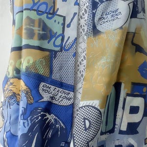 May include: A white and blue scarf with a comic book print. The scarf features a variety of images, including a couple kissing, a cityscape, and the words "Oh I love you!" and "I love you too, David."