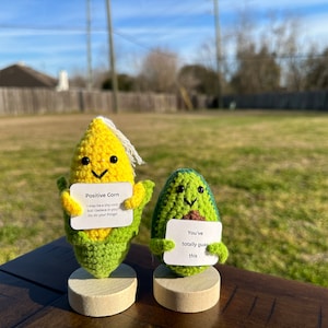 May include: Two crocheted plushies, a corn and an avocado, are holding signs with positive messages. The corn is holding a sign that says "Positive Corn I may be a tiny corn but I believe in you Go do your thing!" The avocado is holding a sign that says "You've totally guac this". The plushies are standing on wooden bases and are in front of a blurred background of a grassy yard.