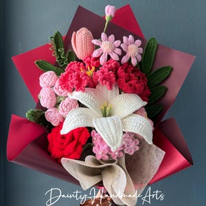 May include: A handmade crochet bouquet featuring a variety of colorful flowers, including red, pink, and white lilies, wrapped in red and cream paper. The bouquet is tied with a brown ribbon that says "Just for you". The text "Dainty Handmade Arts" is also visible.