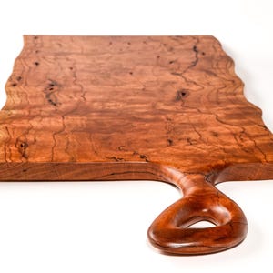 May include: A handcrafted wooden serving board with a natural edge and a handle with a circular opening. The board is made of reddish-brown wood with a wavy edge and a smooth, polished finish. The board is approximately 18 inches long.