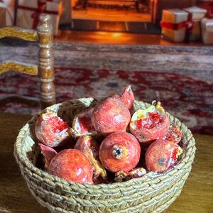 May include: A miniature woven basket filled with red pomegranates, some whole and some cut open to reveal the red seeds. The basket is on a wooden surface, with a blurred background of a fireplace and wrapped gifts.