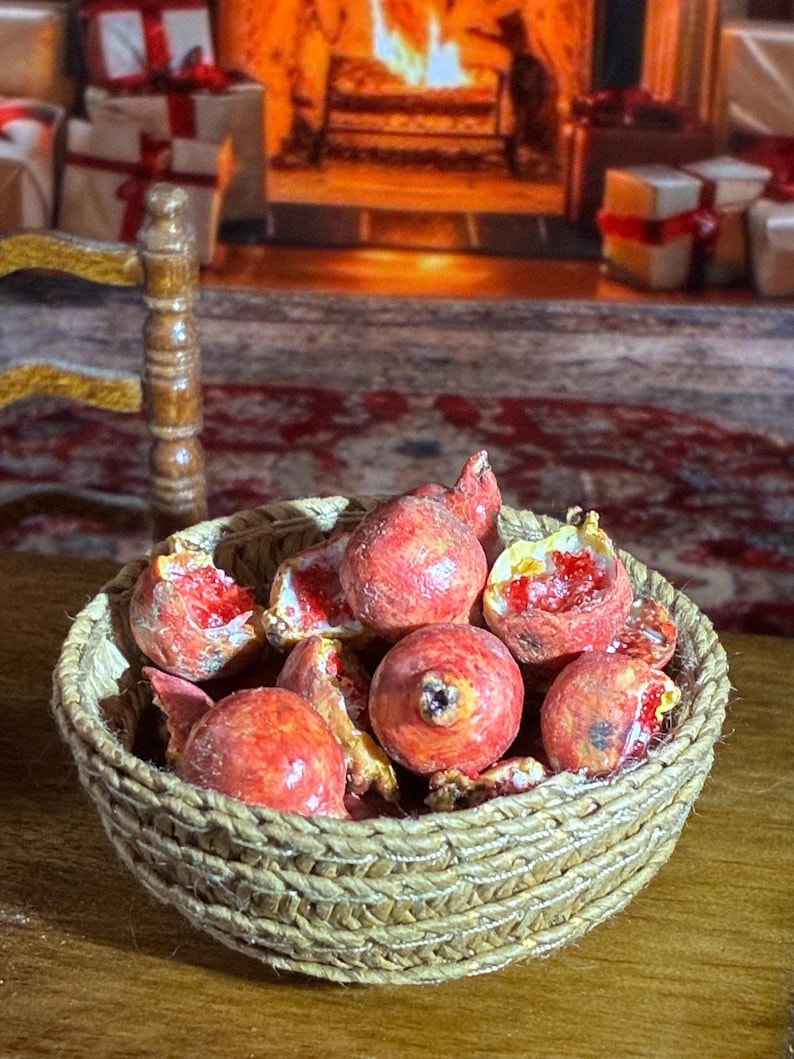 May include: A miniature woven basket filled with red pomegranates, some whole and some cut open to reveal the red seeds. The basket is on a wooden surface, with a blurred background of a fireplace and wrapped gifts.