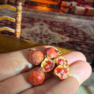 May include: Miniature pomegranates, some whole and some halved, revealing bright red seeds and yellow flesh. The small, detailed fruits are held in a hand, suggesting a dollhouse or miniature food theme. The pomegranates have a realistic, textured appearance.