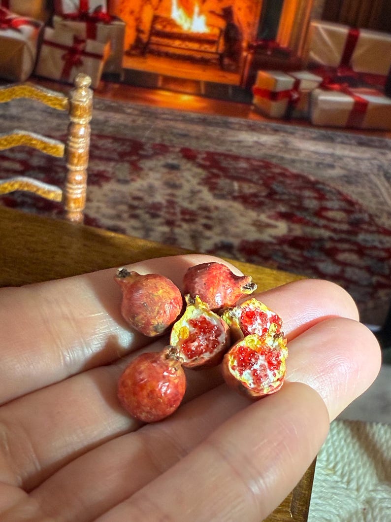 May include: Miniature pomegranates, some whole and some halved, revealing bright red seeds and yellow flesh. The small, detailed fruits are held in a hand, suggesting a dollhouse or miniature food theme. The pomegranates have a realistic, textured appearance.