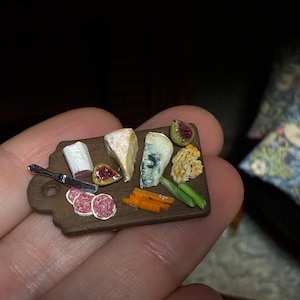 May include: A miniature charcuterie board featuring small replicas of cheese wedges, sliced salami, fig halves, crackers, and green vegetables. A tiny silver-colored knife rests on the brown wooden board, which has a handle with a hole.